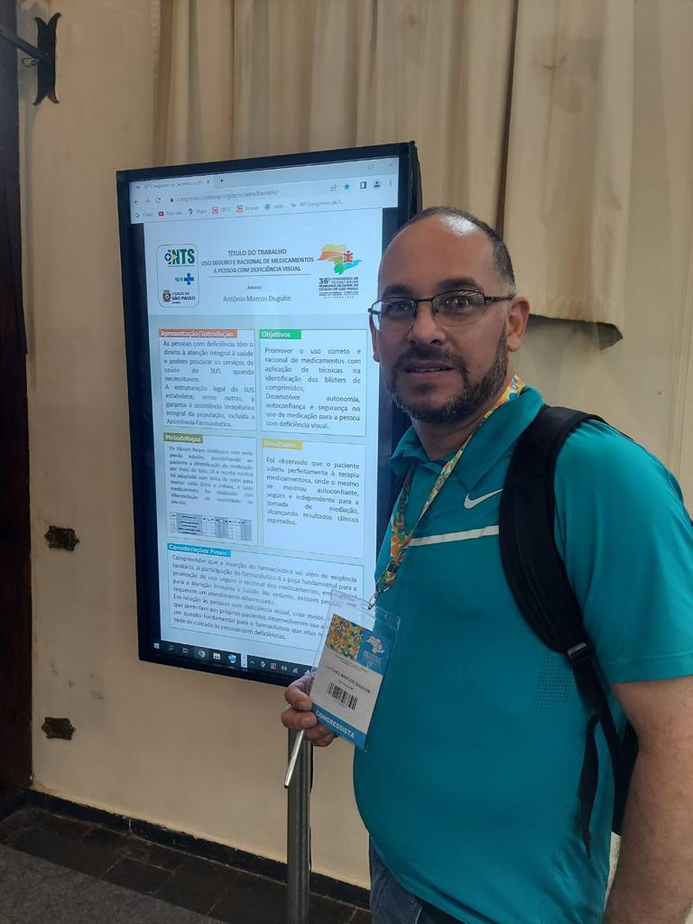 Na imagem, Antônio está ao lado de um painel que traz informações sobre o uso racional de medicamentos. Ele usa óculos, camiseta azul, crachá e mochila.